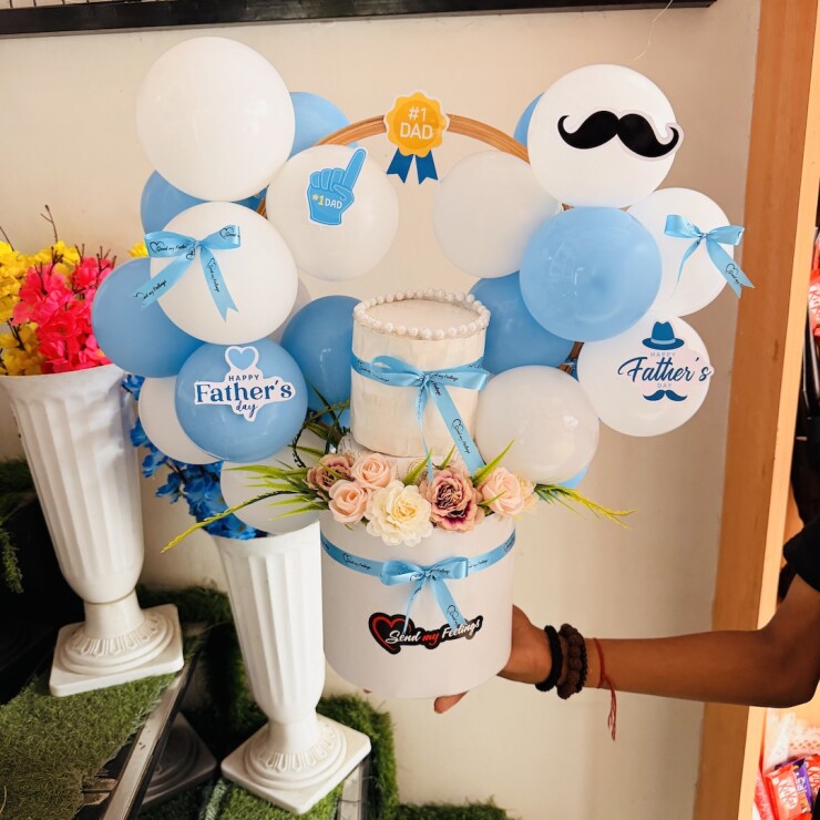 Father's Day gift arrangement with mini pineapple cake, sky blue and white balloons, artificial flowers, and premium white round box by Send My Feelings.
