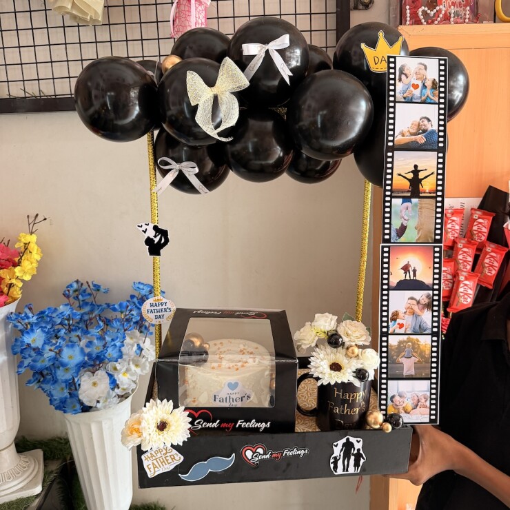 Personalized Father's Day hamper with fruit cake, memory photo strip, floral mug, black balloons, flowers, and themed decorations by Send My Feelings.